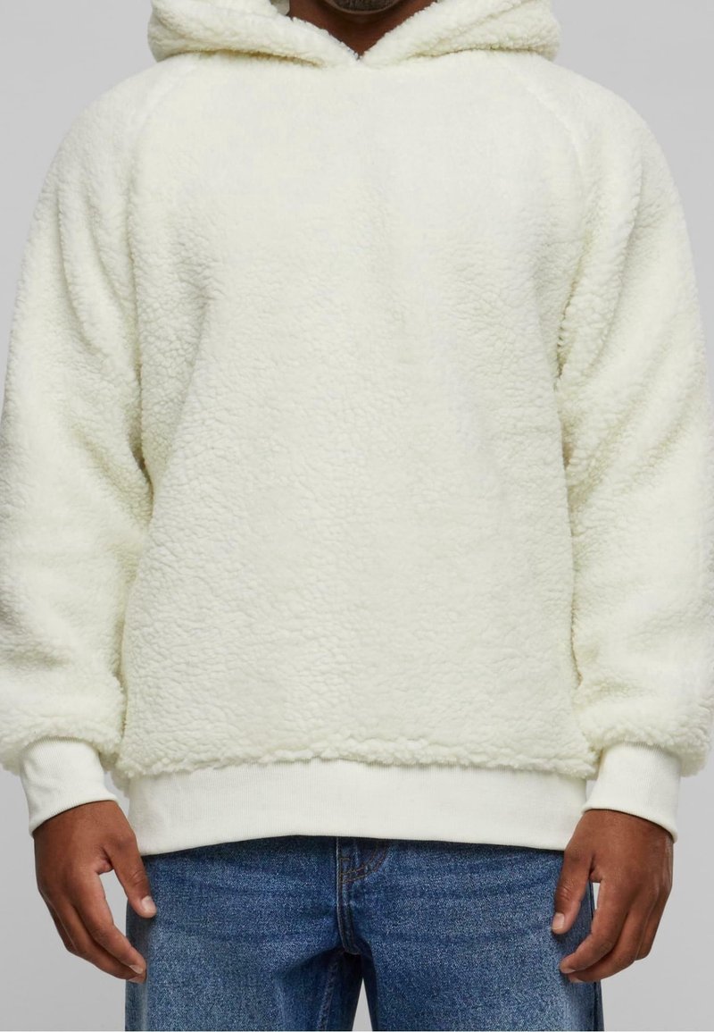 Homme portant un sweat à capuche blanc et duveteux et un jean bleu, debout devant un fond uni.