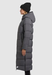 Abrigo acolchado largo en gris oscuro, con capucha, textura acolchada, bolsillos laterales con cremalleras y diseño hasta la altura de la rodilla. Combinado con un gorro negro.