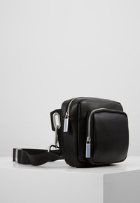 Kompaktowa czarna skórzana torba na ramię z dwoma srebrnymi zamkami błyskawicznymi i regulowanym czarnym paskiem na białej powierzchni.
