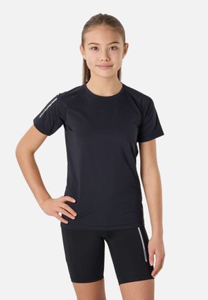 Ung person står med hånden på hoften iført sort kortærmet skjorte og sorte sportsshorts med reflekterende striber på ærmer og ben.