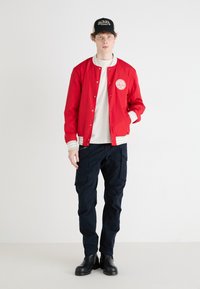 Rote leichte Bomberjacke mit weißen gestreiften Bündchen, darunter ein weißes T-Shirt, dunkle Cargo-Hosen und eine schwarze Kappe mit Logo. Schwarze Stiefel.