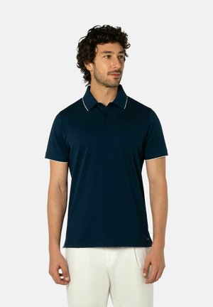 Man draagt een marineblauw poloshirt met witte bies op de kraag en mouwen, gecombineerd met een witte broek, staand tegen een effen achtergrond.