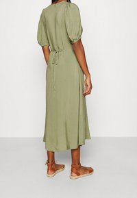 Vihreä oliivinvihreä mekko, jossa on puffihihat ja solmittava vyötärödetalj, midi-pituus ja sileä kangas. Ruskeat hihnatossut täydentävät asun.