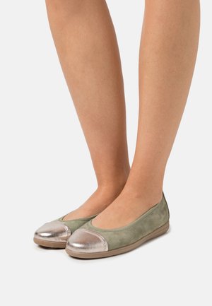 Femme portant des ballerines en daim vert olive avec des embouts métalliques argentés, se tenant sur un fond blanc uni.