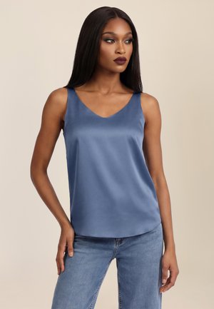 Frau mit langen, glatten Haaren, die ein ärmelloses blaues Satintop und hellblaue Jeans trägt, posiert mit einer Hand in der Hüfte.