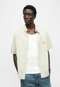 Νέος άνδρας που φοράει ανοιχτοπράσινο πουκάμισο με κοντά μανίκια πάνω από λευκή φανέλα και μπλε τζιν, κοιτάζοντας προς τα αριστερά του.