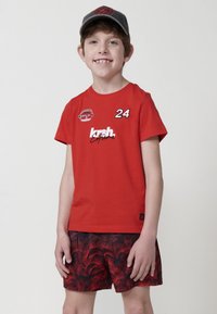 Červené bavlnené tričko s bielym nápisom, číslom 24 a logom. Nosí sa so vzorovanými čiernymi a červenými šortkami.