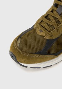 Olive zelena tenisica s mrežastim gornjim dijelom i suede detaljima, crnim naglasima i teksturiranim potplatom. Öice su tkanine i izdržljive.