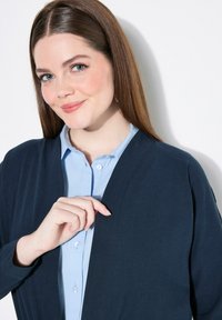 Cardigan blu navy con vestibilità rilassata sopra una camicia azzurro chiaro con bottoni. Il cardigan ha una apertura frontale e una texture liscia.