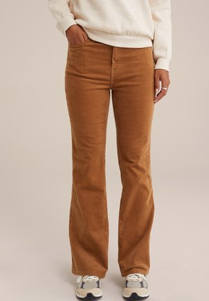 Osoba nosząca brązowe spodnie z wysokim stanem z sztruksu, połączone z beżowo-szarymi sneakersami i jasnokremową bluzą, stojąca z jedną ręką w kieszeni.