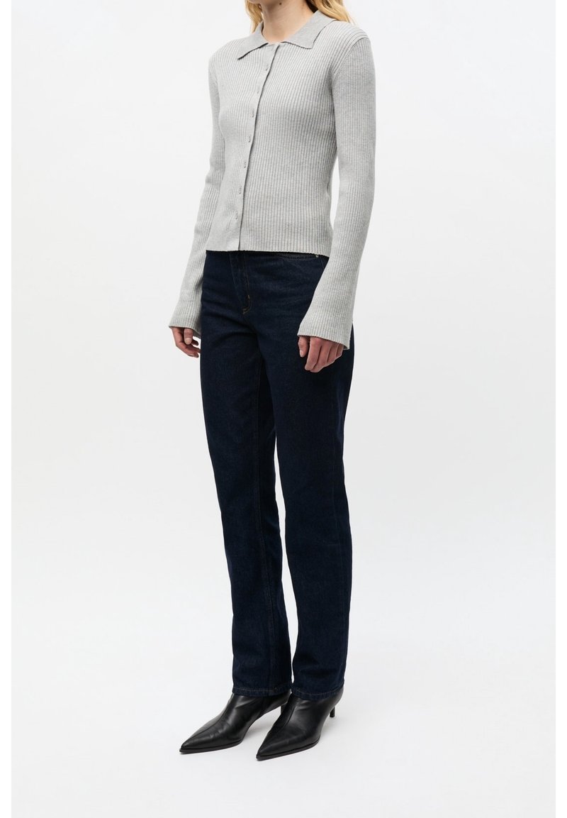 Femme portant un pull boutonné côtelé gris clair, un jean bleu foncé et des bottes à talons noires à bout pointu sur fond blanc.