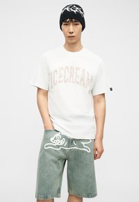 Νεαρός άνδρας που φοράει λευκό μπλουζάκι με την επιγραφή "ICECREAM", τζιν σορτς με λευκό σχέδιο καρτούν σκύλου και μαύρο σκουφάκι με σχέδιο αστραπής.