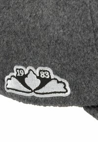 Tissu en laine gris avec un emblème brodé blanc et noir montrant "1983" et un motif stylisé en forme d'aile.