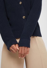 Mörkblå stickad cardigan med en texturerad yta, försedd med träknappar, ribbade ärmslut och en avslappnad passform. Händer håller tyget.