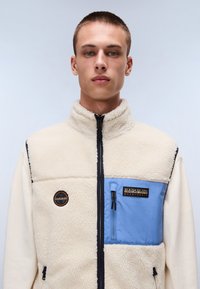 Fleecetröja i krämfärg med en blå nylonbröstficka. Har hög krage, dragkedjestängning och logotypapplikationer. Mjuk textur och kontrasterande detaljer.