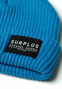 Plava rebrasta pletena kapa s crnom tkaninom na kojoj je ispisano "SURPLUS APPAREL GOODS" i "EST. 1997" bijelim slovima. Mekana tekstura.