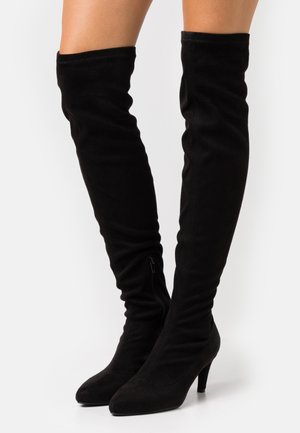 Bottes en daim noir au-dessus du genou avec bouts pointus, talons moyens et fermetures éclair latérales, portées sur des jambes nues sur un fond blanc.