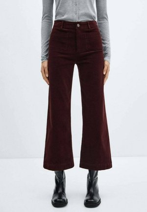 Vrouw die een donkere, wijde, hooggetailleerde broek draagt, een grijze top met lange mouwen en zwarte enkellaarzen, staand tegen een witte achtergrond.