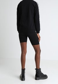 Persona vistiendo pantalones cortos de ciclista negros, sudadera negra con franjas de logo blanco en las mangas y botas de combate negras con cordones, de pie de espaldas.