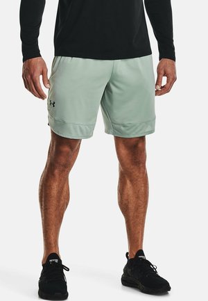 Homme portant un short de sport vert clair avec le logo Under Armour noir, un t-shirt à manches longues noir et des baskets noires, debout sur fond blanc.