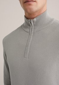 Pulover gri deschis cu guler înalt și fermoar pe jumătate, realizat dintr-un material tricotat texturat cu model în carouri.