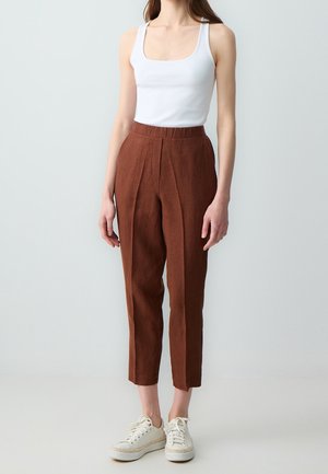 Femme portant un haut blanc sans manches, un pantalon marron court taille haute et des baskets blanches, debout devant un fond uni.