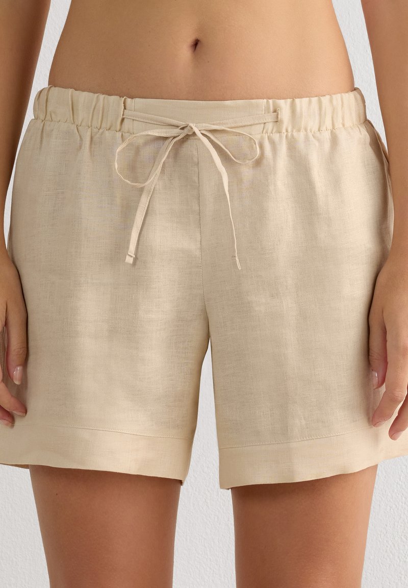Beige Leinen-Shorts mit elastischem Bund und vorne gebundenem Kordelzug, getragen von einer Person mit sichtbarem Bauch und Händen seitlich am Körper.