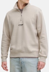 Mężczyzna w beżowym sweatshirtcie z półsuwnym zamkiem z logo "JJ Studios" na piersi oraz w jasnoniebieskich dżinsach, stojący na tle białym.