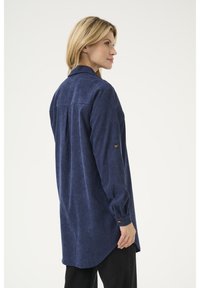 Chemise en velours côtelé bleu marine à manches longues, avec un col et un poignet à boutons. Dispose d'une pliure au dos pour un style et un mouvement supplémentaires.