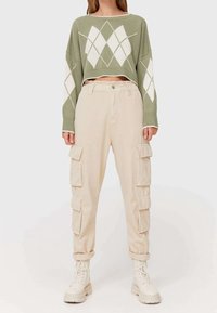 Pull en tricot vert coupé avec un motif de losanges blancs, associé à un pantalon cargo beige avec plusieurs poches et des bottes blanches à lacets.