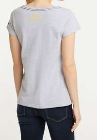 Gray short-sleeve T-shirt with a rounded neckline and yellow logo detail on the back, worn with dark blue jeans, showing a casual fit.