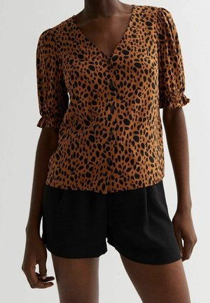 Žena nosící hnědou košili s leopardím potiskem a knoflíky, s nafouklými rukávy a černé krátké kalhoty s vysokým pasem.