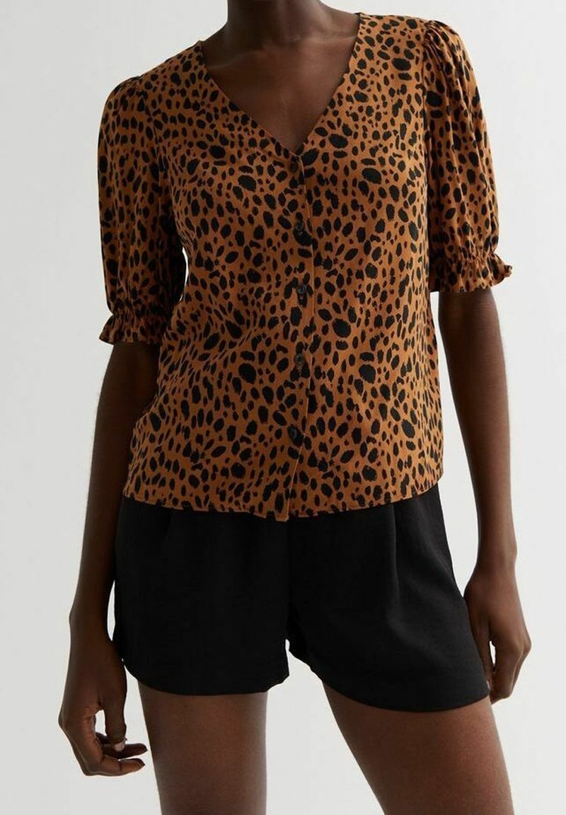 Femme portant un chemisier marron imprimé léopard avec boutons, manches bouffantes et un short noir taille haute.