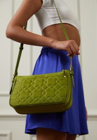 Borsa a tracolla verde trapuntata con una texture liscia, dotata di una tracolla e hardware in tonalità oro. Mostrata con una gonna blu in un ambiente neutro.