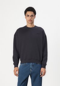 Mørkegrå hettegenser med rund hals, oppbrettede lange ermer og en diskret logo på brystet. Kombinert med blå jeans. Glatt tekstur.