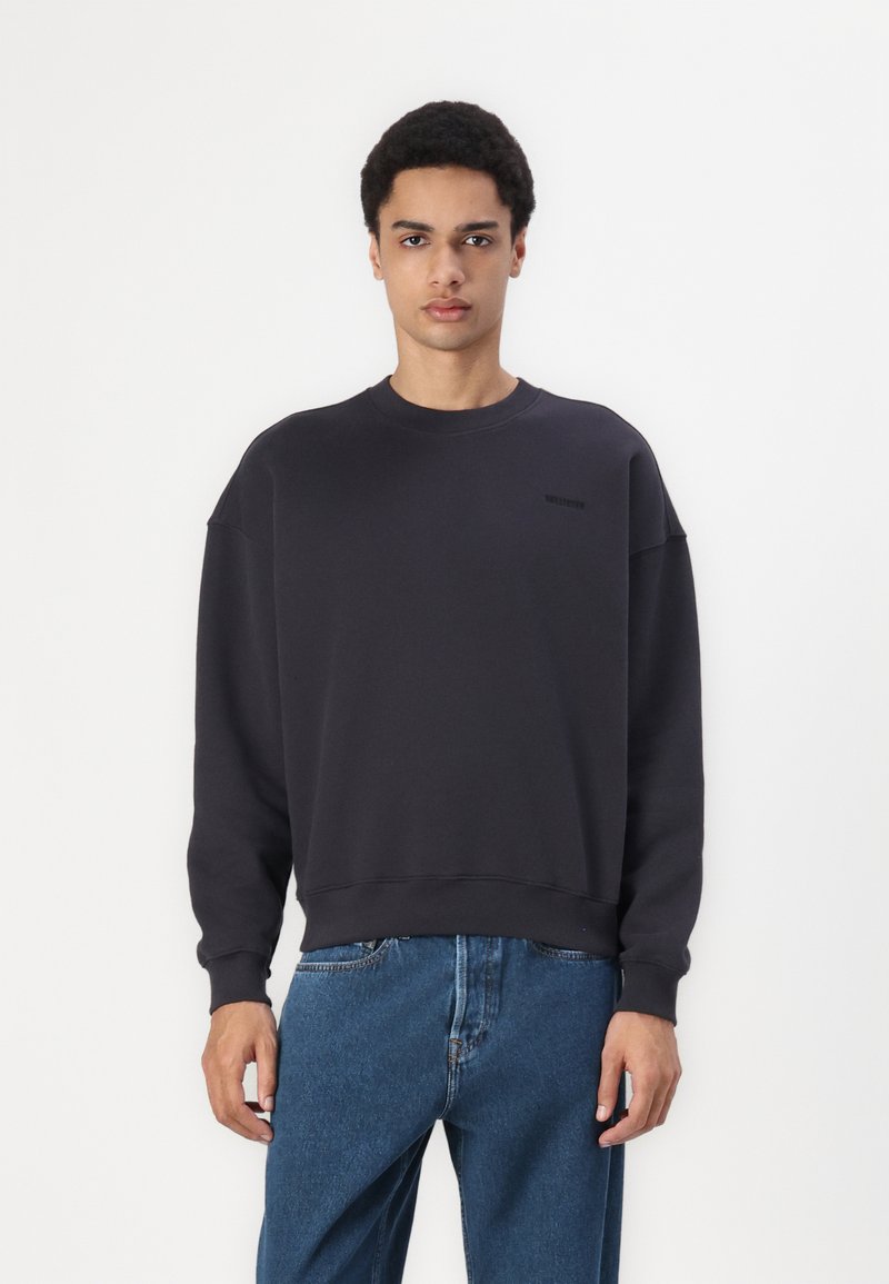 Mørkegrå hettegenser med rund hals, oppbrettede lange ermer og en diskret logo på brystet. Kombinert med blå jeans. Glatt tekstur.