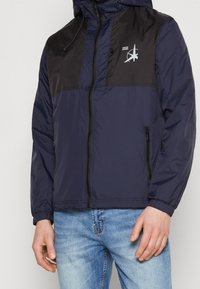 Giacca in nylon blu navy e nera con chiusura a zip, cappuccio e due tasche laterali. Logo stampato sul petto, tessuto testurizzato, polsini elastici.