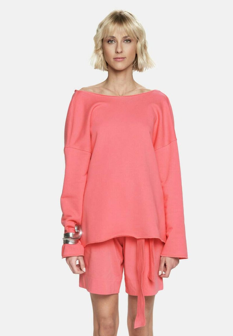 Przesadnie duży, jasnoróżowy sweter z szerokim dekoltem i długimi rękawami, w zestawie z dopasowanymi szortami. Srebrny dodatek na lewym nadgarstku.