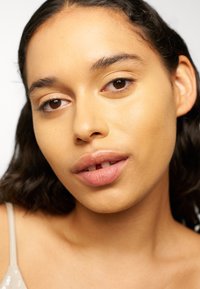 Primer plano de la cara de una persona con piel marrón media, cabello oscuro y ondulado, maquillaje natural, labios ligeramente separados, mostrando un espacio entre los dientes.