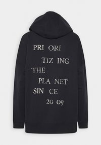 Černá mikina s kapucí s opotřebovaným bílým nápisem na zádech "Prioritizujeme planetu od roku 2009."
