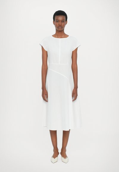 Robe blanche avec un corsage ajusté et une jupe évasée. Manches courtes et détails de couture en diagonale. Associée à des chaussures blanches à talons pointus.