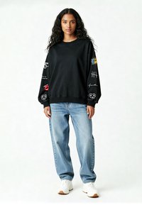 Felpa nera con stampe grafiche sulle maniche, abbinata a jeans baggy azzurri. Sneakers bianche completano l'outfit casual.