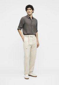 Ένας άνδρας στέκεται με τα χέρια στις τσέπες, φορώντας σκούρο γκρι πουκάμισο με κουμπιά και γυρισμένα μανίκια, κρεμ ψηλόμεσο παντελόνι με ζώνη, λευκά αθλητικά παπούτσια.