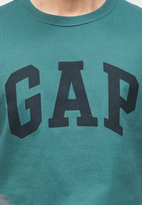 Κοντινό πλάνο σε μπλουζάκι τυρκουάζ με μεγάλα μαύρα γράμματα "GAP" τυπωμένα στο μπροστινό μέρος, που φοράει ένα άτομο.