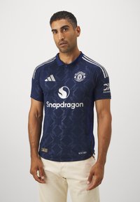 Námořnická modrá dres Manchester United s límečkem, s texturovaným vzorem, bílými logy a třemi bílými pruhy na ramenou.
