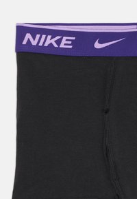 Crne atletske kratke hlače s ljubičastim pojasom na kojem je bijeli tekst i logo "NIKE", glatke teksture i ojačanim šavovima.