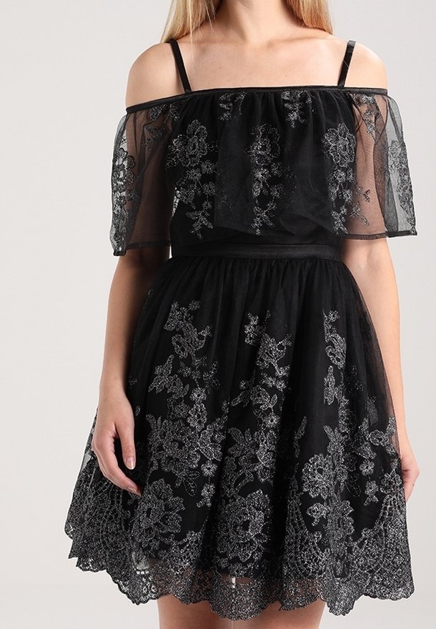 Femme portant une robe noire épaules dénudées avec broderie florale argentée et superposition de dentelle transparente sur les manches et la jupe.