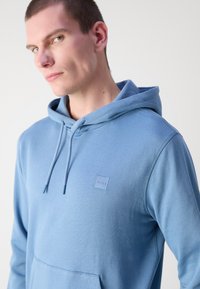 Mau de capuz azul claro feito de algodão macio, com um bolso canguru e um retângulo de logótipo no peito. Cordões no capuz.