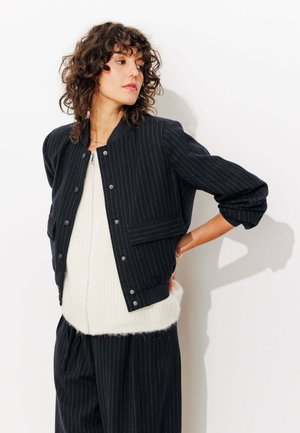 Veste bomber rayée marine avec fermetures à bouton et poches à rabat, associée à un pull crème. Texture douce, coupe décontractée et poignets côtelés.