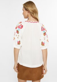 Bluză albă cu broderie florală colorată pe mânecile bufante, material texturat și un guler roz cu margine ondulată. Asortată cu o fustă din suede maro.
